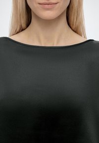Femme aux cheveux blonds raides portant un haut noir simple à encolure bateau, montrée des épaules à la bouche sur un fond clair.