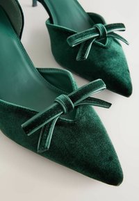 Pantofi cu toc înalt din catifea verde, cu vârf ascuțit, evidențiați de un accent cu fundiță în față și o căptușeală interioară netedă.