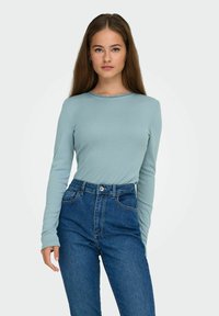 Langermet lyseblå topp med bølgete halslinning, kombinert med høytlivs blå denimbukser som viser en jevn tekstur.