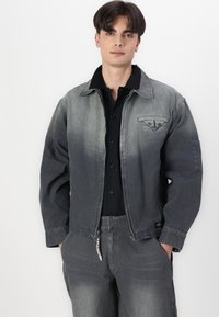 Gray denim jacket with an ombre effect, featuring a chest pocket, zipper closure, and visible stitching. Paired with a black button-up shirt.