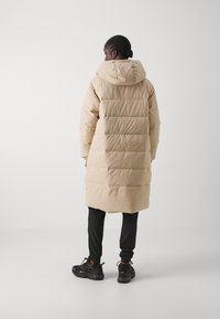 Veste doudoune beige avec une capuche, présentant des motifs matelassés horizontaux. Portée avec un pantalon de jogging noir et des baskets noires texturées.