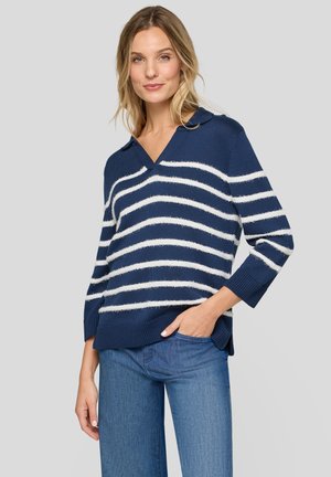 Woman wearing a navy blue sweater with white horizontal stripes and blue jeans, standing with one hand in her pocket against a plain background.