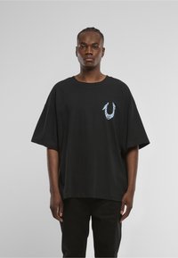 Czarny, oversize'owy T-shirt z bawełny z okrągłym dekoltem. Z przodu na wysokości klatki piersiowej znajduje się niebiesko-biały wzór graficzny.