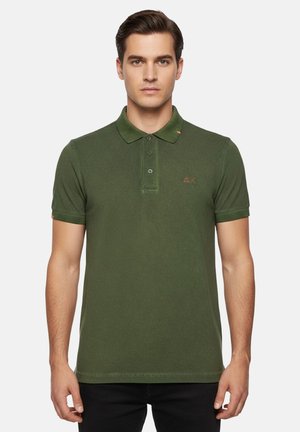 Uomo che indossa una polo verde scuro aderente a maniche corte con abbottonatura a tre bottoni, in piedi contro uno sfondo neutro.