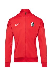 Rode sportjack met een volledige rits, gemaakt van gladde stof, met een wit Nike-logo en een zwart-wit teamembleem op de borst.