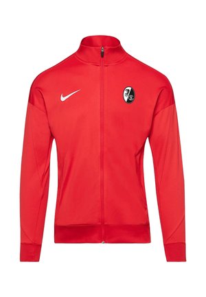 Rote Sportjacke mit vollem Reißverschluss, aus glattem Stoff, mit einem weißen Nike-Logo und einem schwarz-weißen Teamwappen auf der Brust.