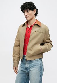 Jeune homme aux cheveux bouclés portant une veste beige avec un col marron, une chemise rouge boutonnée et un jean bleu clair, debout devant un fond uni.