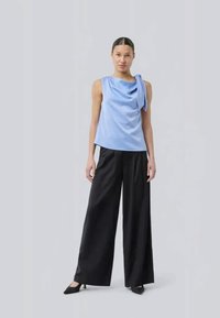 Top sem mangas azul claro com um design drapeado no ombro, combinado com calças largas pretas e sapatos pretos de bico.