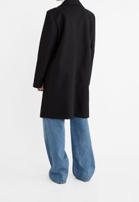 Persoon met een lange zwarte jas en losse blauwe jeans die tegen een effen witte achtergrond staat, gezien van achteren.