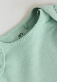 Svijetlozeleni pamučni top bez rukava s rebrastim izrezom i vidljivom crnom polukružnom oznakom unutar ovratnika.