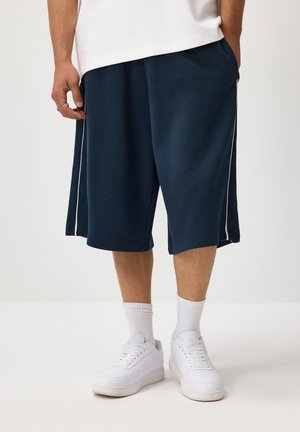 Man wearing navy blue knee-length shorts, white socks, white sneakers, and a white shirt, standing with hands in pockets.