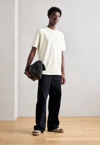 Tricou crem unisex cu mânecă scurtă, pantaloni negri lejeri, ținând o geantă din piele neagră. Podea din lemn și fundal neutru.