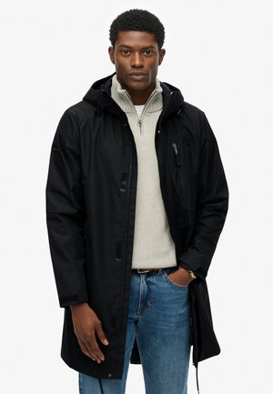 Sort vandtæt jakke med hætte, der har en lynlåslomme på brystet, justerbare manchetter og en afslappet pasform over en lys grå sweater.