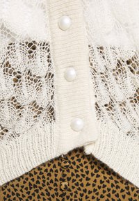 Krémový pletený kardigan s krajkovými detaily a perlami, vrstvený přes hnědý oděv s černým vzorem srdce.