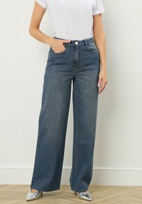 Jean en denim taille haute avec une coupe évasée, présentant un lavage bleu foncé, de subtils clous argentés et des poches avant. Associé à des talons argentés.