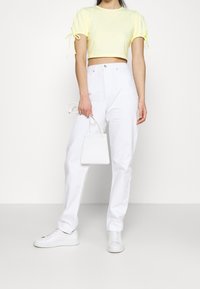 Lysegul kort top med korte puffærmer og bindedetaljer, kombineret med hvide bukser med høj talje og en lille hvid håndtaske.