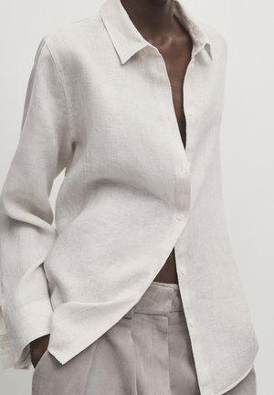 Unbuttoned off-white linen shirt with long sleeves worn tucked into high-waisted light gray trousers.