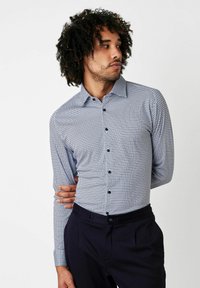 Langärmliges Hemd mit blau-weißer Musterung, Knopfkragen und schwarzen Knöpfen, getragen mit dunklen Hosen. Glattes Material, taillierte Passform.