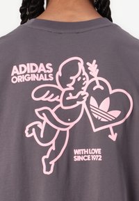 Pilkas marškinėliai su rožiniu grafiniu atvaizdu, kuriame yra cherubas, laikantis širdį su Adidas logotipu. Tekstas skamba: "ADIDAS ORIGINALS SU MEILE NULAT 1972."