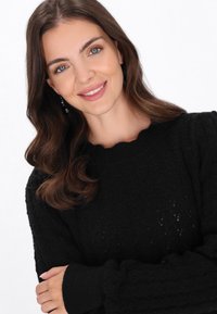 Czarny sweter dzianinowy z teksturowanym wzorem, z koronkowym dekoltem i długimi rękawami. Model pozuje z rękami skrzyżowanymi.