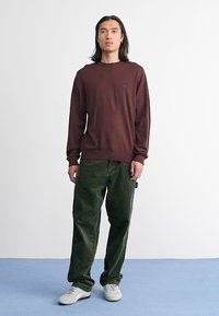 Brauner Langarmpullover mit Rundhalsausschnitt; grüne Cordhosen mit strukturierten Streifen; weiße Sneakers; Modell steht vor einem schlichten Hintergrund.