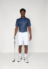 Sötétkék kockás galléros póló rövid ujjú, fehér rövidnadrág és fehér öv. Kiegészítők: golfütő és teniszcipő.