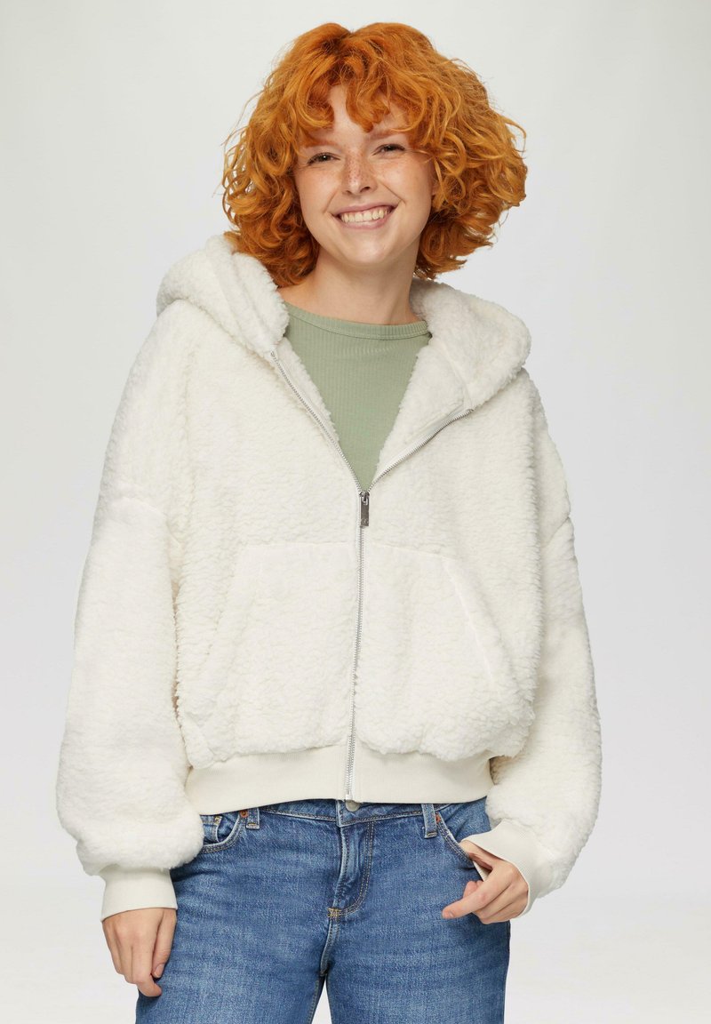 Weiße Fleece-Zip-Hoodie mit Kapuze, großen Fronttaschen und gerippten Bündchen. Über einem hellgrünen Shirt getragen und mit blauen Jeans kombiniert.