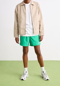 Beige lättviktsjacka över en vit T-shirt, i kombination med gröna shorts och vita sneakers. Vita strumpor synliga ovanför ankeln.