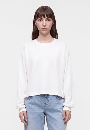 Mujer con cabello castaño liso, vestida con una camiseta blanca de manga larga sencilla y vaqueros azul claro, de pie frente a un fondo claro y liso.
