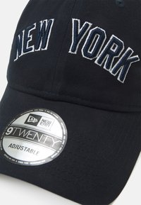 Marineblaues Cap mit "NEW YORK" in Weiß und Blau gestickt. Mit gebogenem Schirm und verstellbarem Riemen. Enthält ein silbernes Größenetikett.