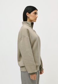 Kvinne med rett svart hår iført en beige zip-up hettegenser og rutete bukser, står i profil mot en enkel hvit bakgrunn.