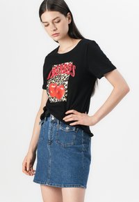 Svart bomulls-t-shirt med röd text "Cherries" och en grafisk bild av två röda körsbär på en leopardmönstrad bakgrund, kombinerad med en jeanskjol.
