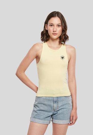 Femme aux cheveux bruns courts porte un haut sans manches jaune pâle avec un motif cœur noir et un short en jean bleu clair retroussé.