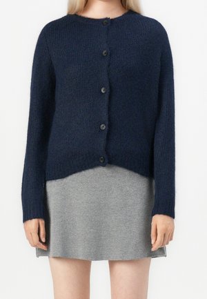 Femme portant un cardigan bleu marine boutonné sur une jupe courte grise, debout devant un fond uni.