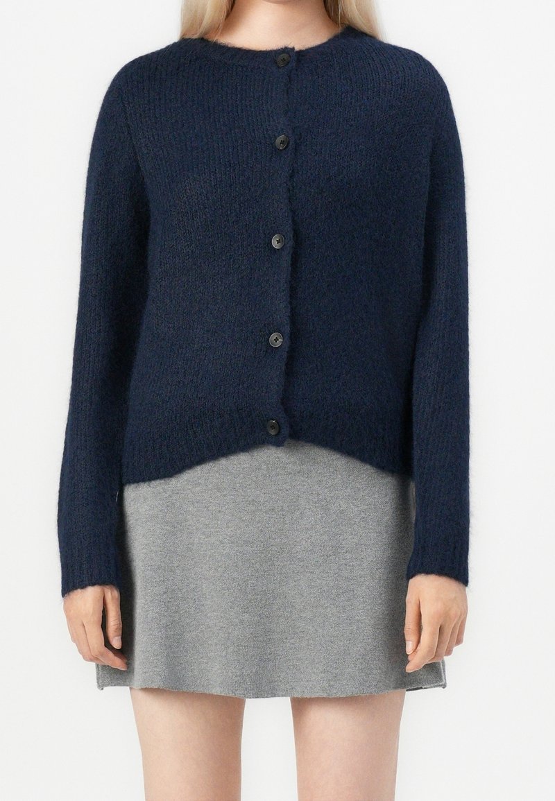 Femme portant un cardigan bleu marine boutonné sur une jupe courte grise, debout devant un fond uni.