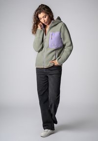 Fleecetak in het groen met een paars borstzakje, capuchon, ritssluiting en opvallende naden. Draagt zwarte jeans en witte sneakers.