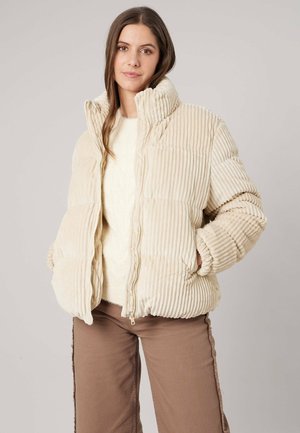 Vrouw met lang bruin haar die een crèmekleurige geribde donzen jas, een witte trui en bruine broek draagt, staand met handen in de jaszakken.