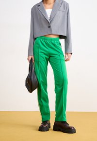 Blazer gris recortado sobre una blusa blanca, combinado con pantalones deportivos verdes con franjas laterales y botas negras gruesas. Sosteniendo un bolso negro.