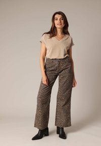 Femme debout, les mains détendues, portant un haut beige à col en V, un pantalon large à imprimé léopard et des bottines à talons noires sur un fond neutre.