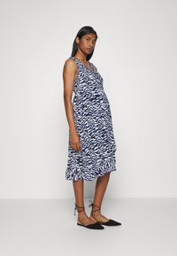 Vestido de maternidade sem mangas em azul-marinho com padrão abstrato branco, com bainha de babados e decote em V. Combinado com sapatos pretos planos.