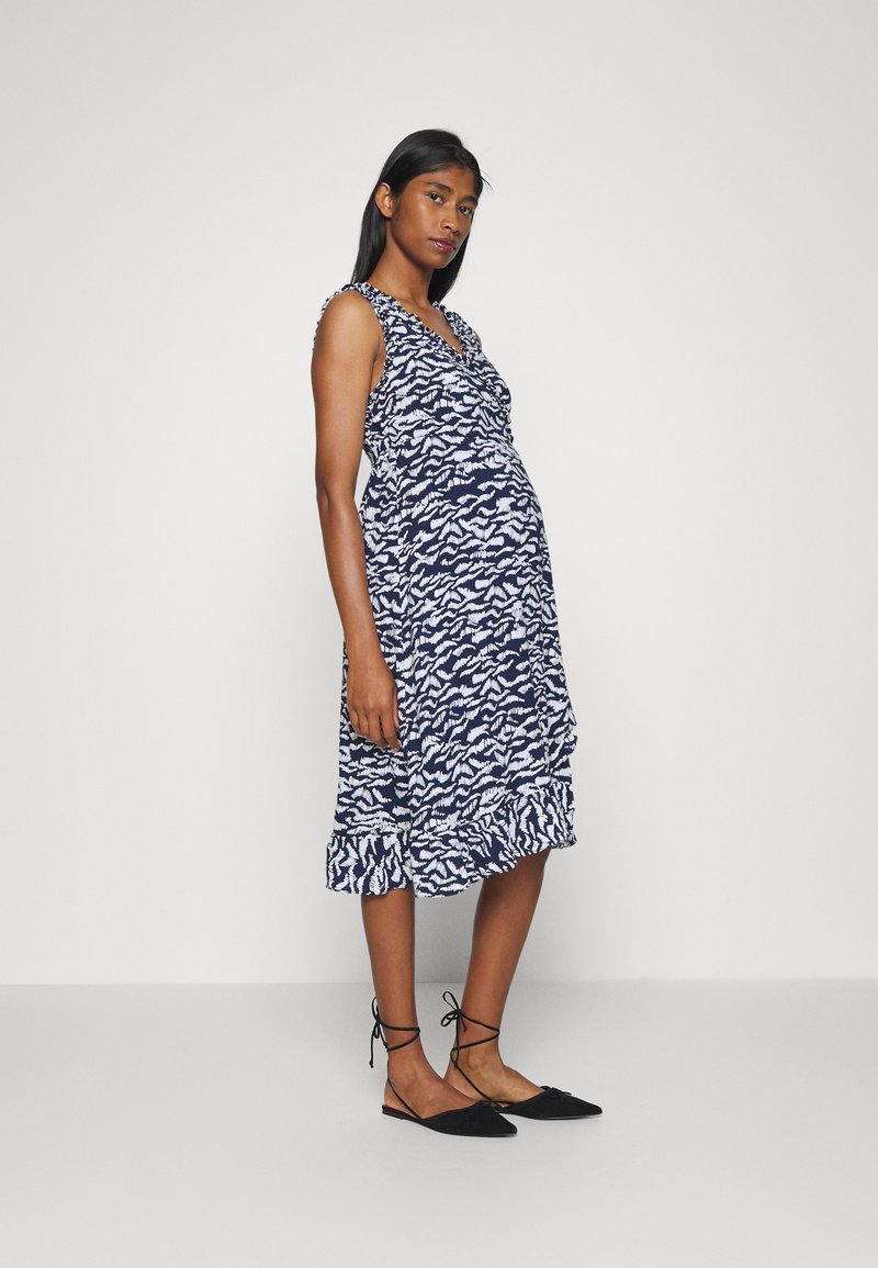 Vestido de maternidade sem mangas em azul-marinho com padrão abstrato branco, com bainha de babados e decote em V. Combinado com sapatos pretos planos.