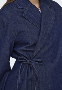 Tume sinine denim jakk, millel on ümberpööratud disain, sakilised kaelused ja sidumisuskumuse sulgemine vööl. Toodud on kontrastselt õmmeldud detail.
