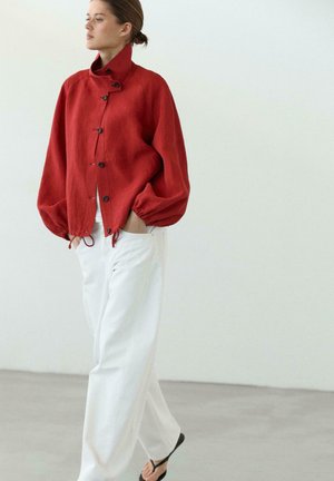 Frau trägt eine lockere rote Jacke mit Knöpfen, weiße Hose und schwarze Sandalen, steht mit den Händen in den Taschen vor einem einfarbigen hellen Hintergrund.