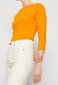 Langes oranges Oberteil mit einem Bootsausschnitt, kombiniert mit weißen hochtaillierten Jeans. Auffälliges Markenlogo auf der Brust. Glatte Textur.