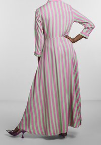 Robe maxi avec des rayures verticales en rose et vert. Manches longues, col et tissu fluide avec une taille marquée. Escarpins violets.