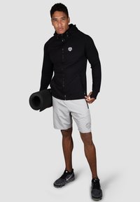 Schwarze Zip-Hoodie mit Logo, graue Shorts mit Muster und schwarze Sportschuhe. Hält eine Rolle einer schwarzen Yogamatte.