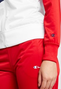 Gros plan d'une personne portant un pantalon de jogging rouge Champion et une veste blanche zippée avec des manches rouges, montrant les logos Champion sur le pantalon et la manche.