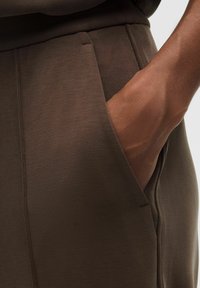 Pantalones marrones con una textura suave y un bolsillo lateral, con una mano parcialmente visible en el interior. Presentan líneas limpias y un diseño minimalista.