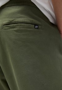 Pantalones de color verde oliva hechos de una tela suave, con un bolsillo trasero que presenta una etiqueta de logo negra. Textura suave y diseño minimalista.