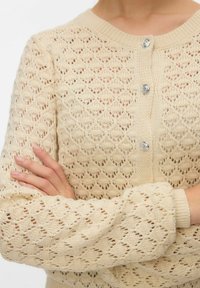 Cardigan beige en tricot avec motif ajouré en losanges, encolure ronde et trois boutons décoratifs. Poignets et ourlet côtelés ajoutant de la texture.